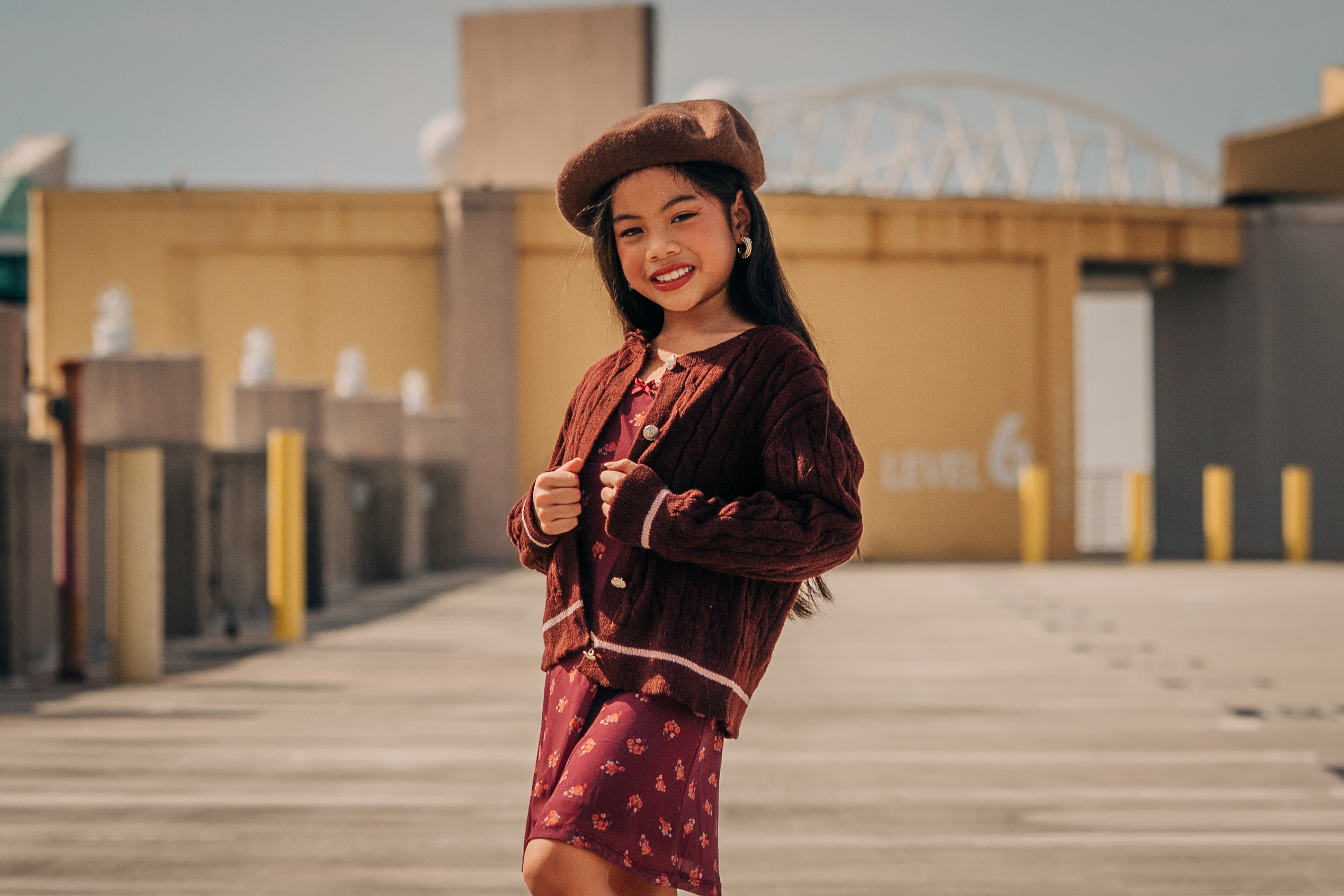 Smiling child model Pichara Luong in a brown beret and burgundy cable-knit cardigan during an outdoor rooftop fashion shoot